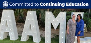 Two smiling women pose together outdoors in front of large illuminated marquee letters spelling “AAML.” One woman wears a patterned dress and the other a vibrant blue dress. Palm trees and lush greenery are visible in the background.